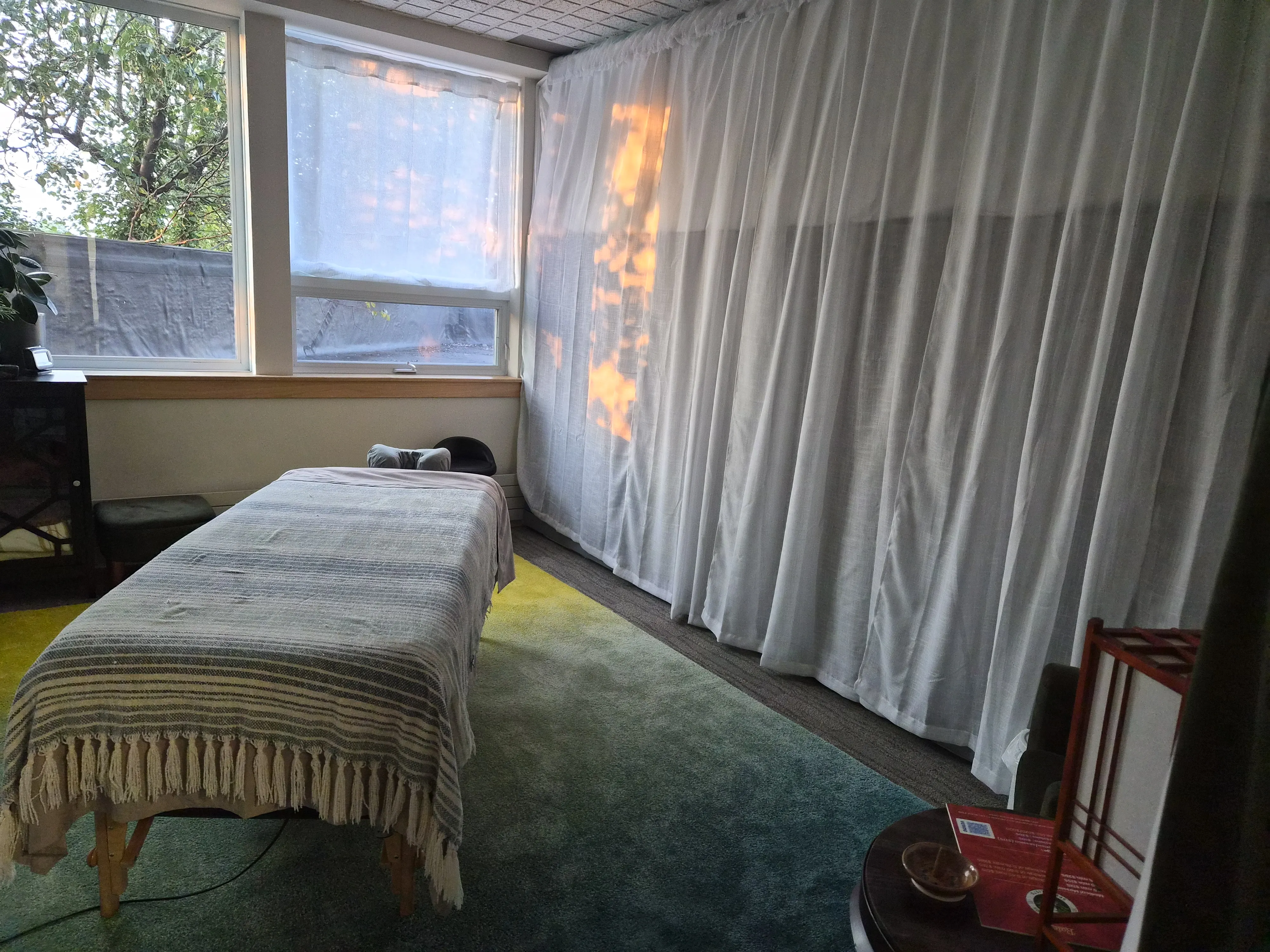 A corner of the treatment room with the massage table beside sheer floor-length curtains and late afternoon light